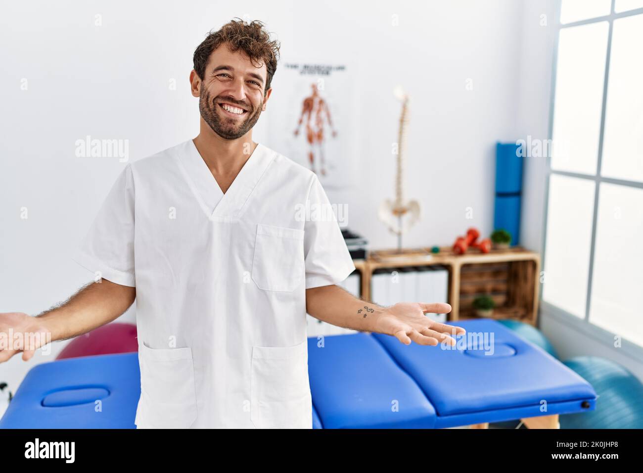 Jeune beau physiothérapeute homme travaillant à la clinique de rétablissement de la douleur souriant gai avec les bras ouverts comme accueil amical, positif et confiant greetin Banque D'Images