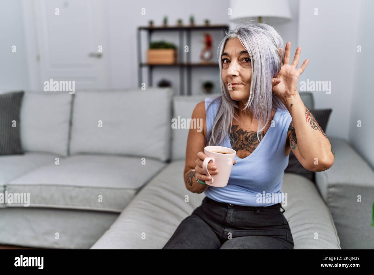 Femme d'âge moyen à cheveux gris buvant du café assis sur le canapé à la maison en souriant avec la main sur l'oreille écoutant une audition de rumeurs ou de potins. Surdité Banque D'Images