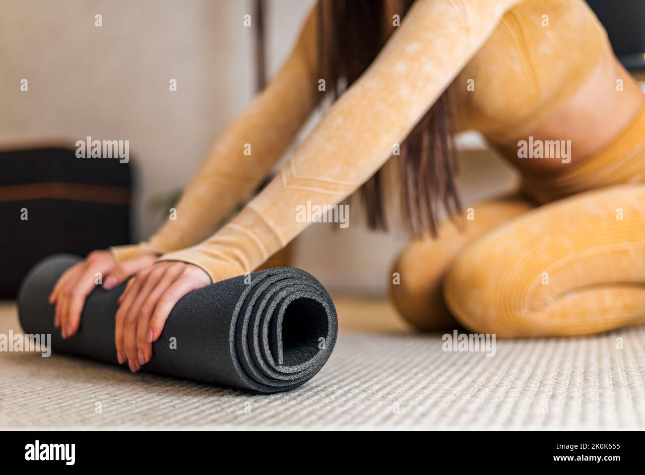 Photo d'une jeune femme brunette énergique, portant un survêtement jaune, pliant son tapis de yoga noir. Banque D'Images