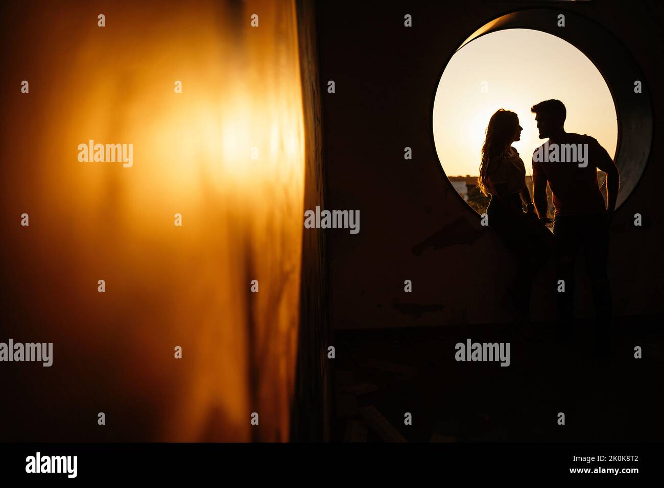 Anonyme couple silhouette dans des vêtements décontractés regardant les uns les autres et rire en s'asseyant à côté de trou de fenêtre rond de bâtiment moderne contre le blu Banque D'Images