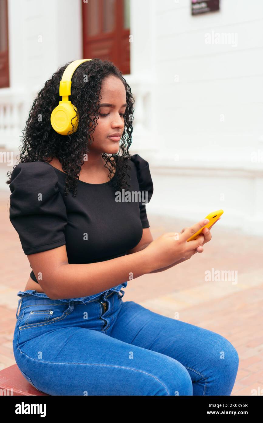Femme ethnique gaie aux cheveux bouclés, écoutant de la musique dans un casque jaune et parcourant un smartphone dans la rue Banque D'Images