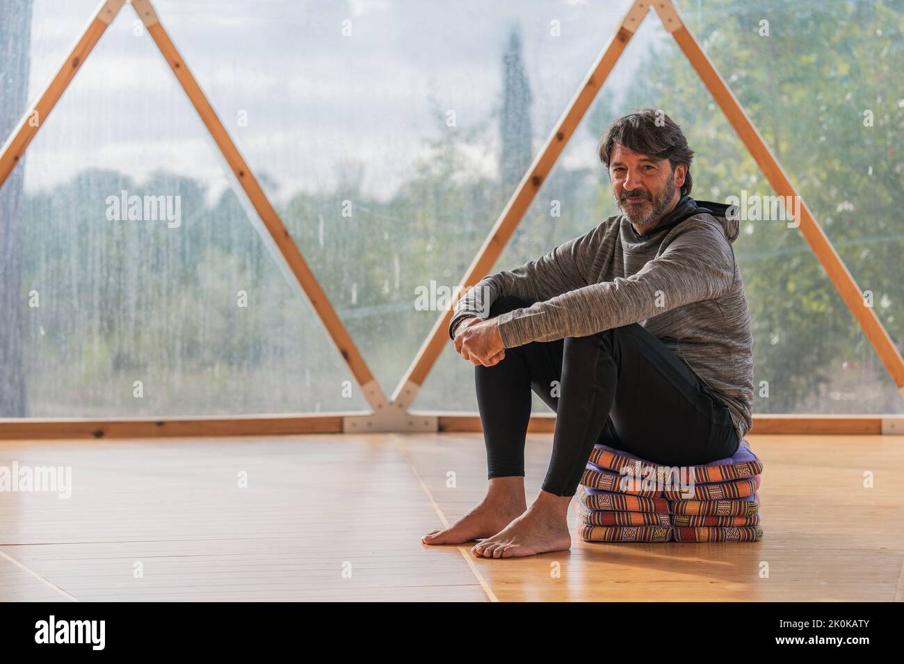 Vue latérale d'un homme gris à poils longs et joyeux, pieds nus et menton reposant sur une main, assis sur des couvertures pliées dans une pièce en bois, à proximité d'une vue panoramique Banque D'Images