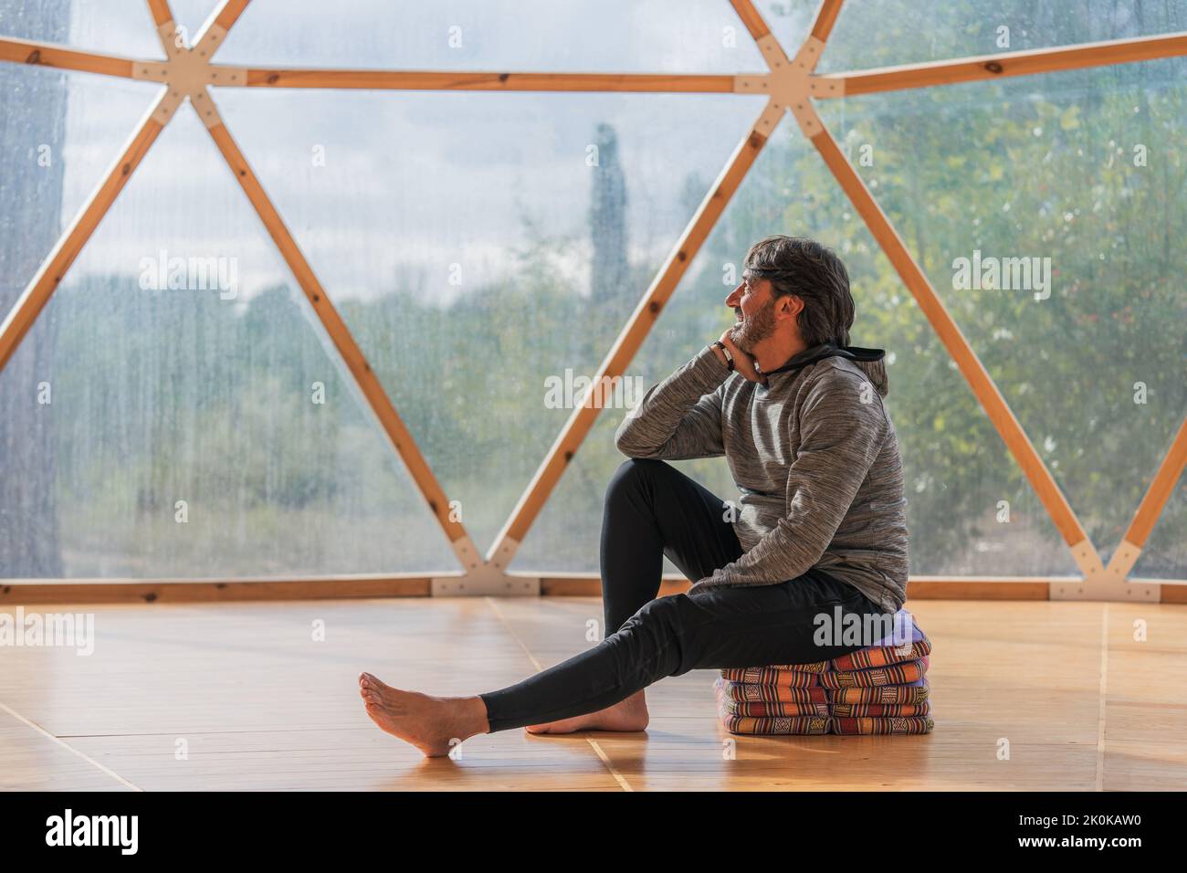 Vue latérale d'un homme gris à poils longs et joyeux, pieds nus et menton reposant sur une main, assis sur des couvertures pliées dans une pièce en bois, à proximité d'une vue panoramique Banque D'Images