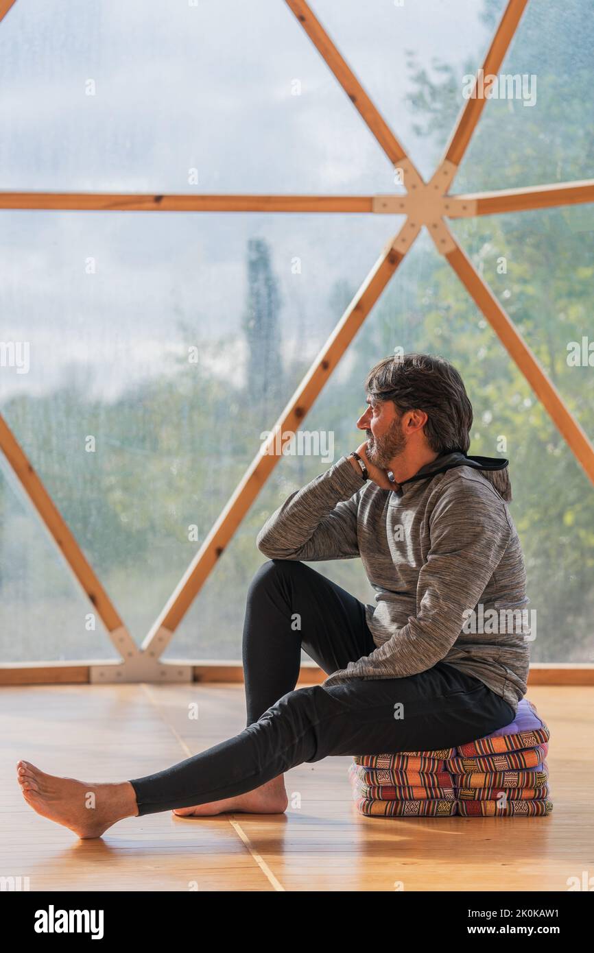 Vue latérale d'un homme gris à poils longs et joyeux, pieds nus et menton reposant sur une main, assis sur des couvertures pliées dans une pièce en bois, à proximité d'une vue panoramique Banque D'Images