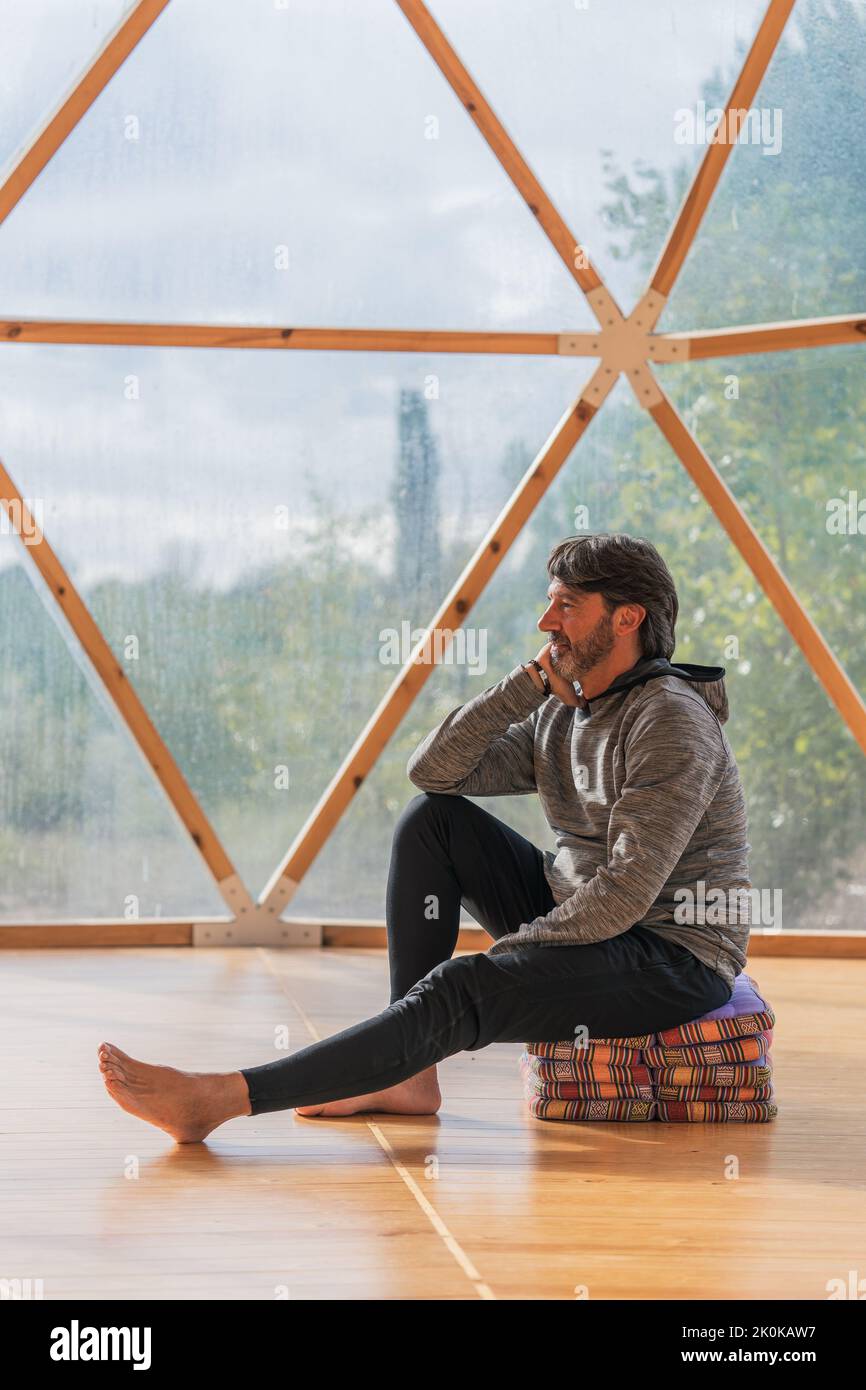 Vue latérale d'un homme gris à poils longs et joyeux, pieds nus et menton reposant sur une main, assis sur des couvertures pliées dans une pièce en bois, à proximité d'une vue panoramique Banque D'Images