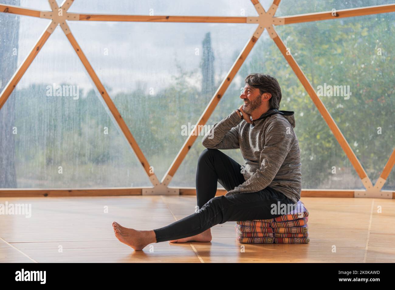 Vue latérale d'un homme gris à poils longs et joyeux, pieds nus et menton reposant sur une main, assis sur des couvertures pliées dans une pièce en bois, à proximité d'une vue panoramique Banque D'Images