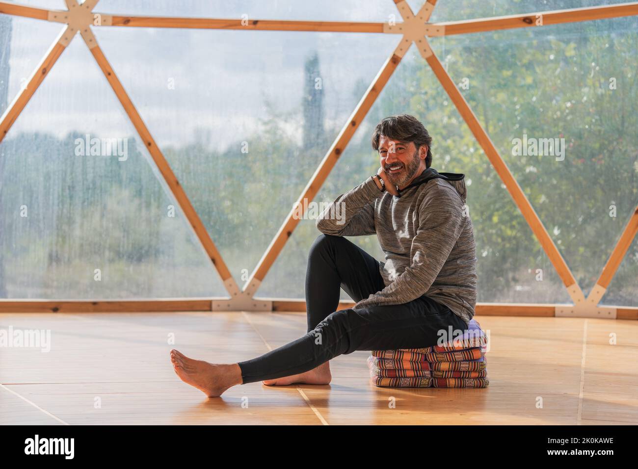 Vue latérale d'un homme gris à poils longs et joyeux, pieds nus et menton reposant sur une main, assis sur des couvertures pliées dans une pièce en bois, à proximité d'une vue panoramique Banque D'Images