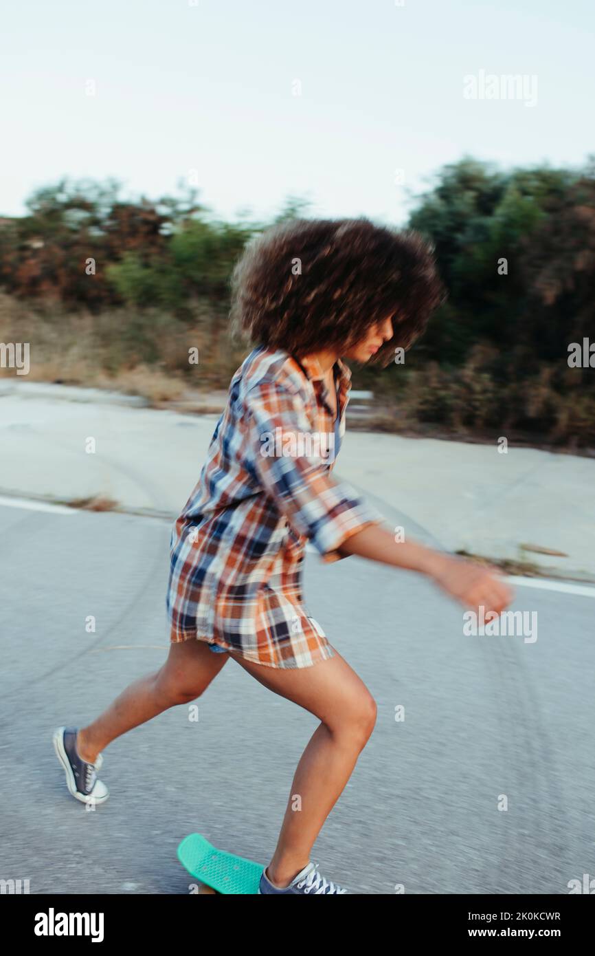 Vue latérale de l'hipster féminin avec la coiffure afro équitation panneau de penny le long de la route vide pendant le coucher du soleil Banque D'Images