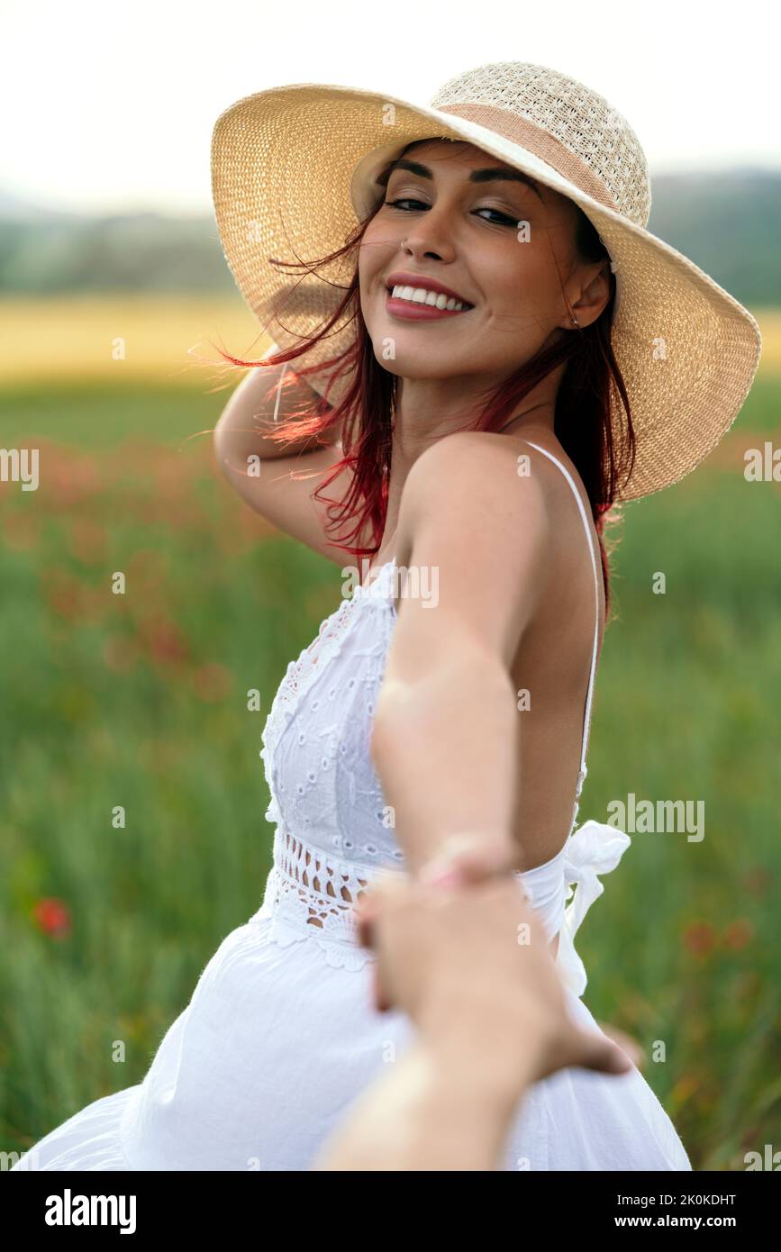 Vue latérale de la femme gaie dans la sundress et chapeau de paille tenant la récolte anonyme partenaire à la main tout en regardant l'appareil photo sur la prairie Banque D'Images