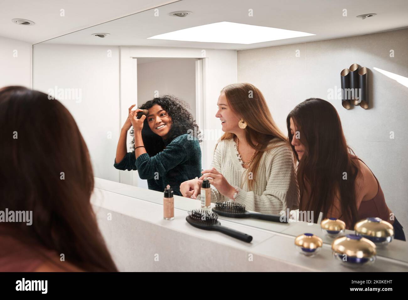 Joyeux jeunes femmes multiethniques amis en tenue décontractée appliquant le maquillage et bavardant contre le miroir dans le dressing ou la salle de bains Banque D'Images