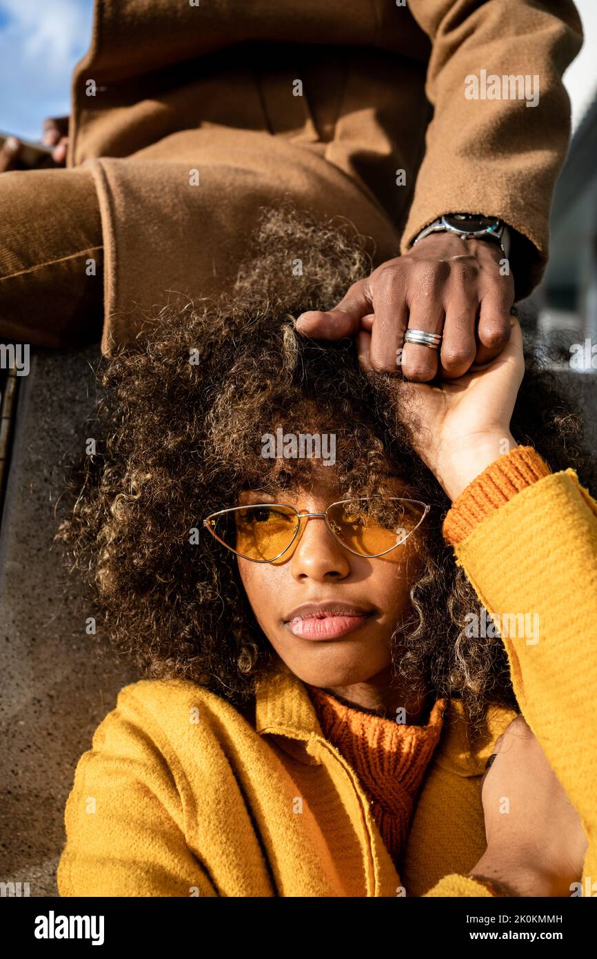 Jeune femme ethnique élégante avec des boucles afro portant des lunettes de soleil tendance et un manteau jaune tenant la main de crop boyfriend à la lumière du soleil Banque D'Images