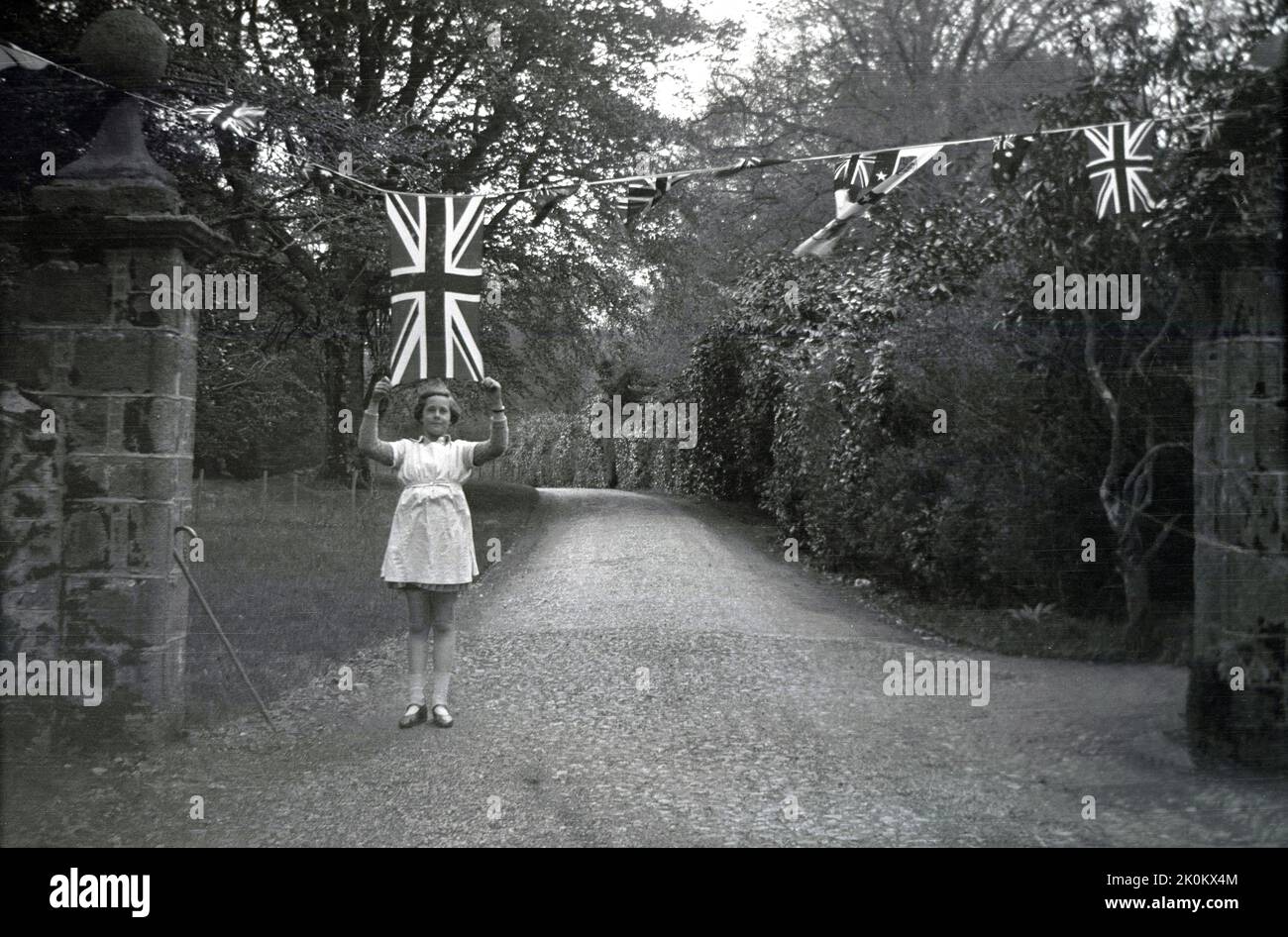1937, historique, à une entrée de la propriété de Forcet Hall, North Yorkshire, Angleterre, Royaume-Uni, une jeune fille tenant un drapeau de l'Union Jack sur le bandeau aérien, célébrant le couronnement de George VI en tant que roi. Banque D'Images