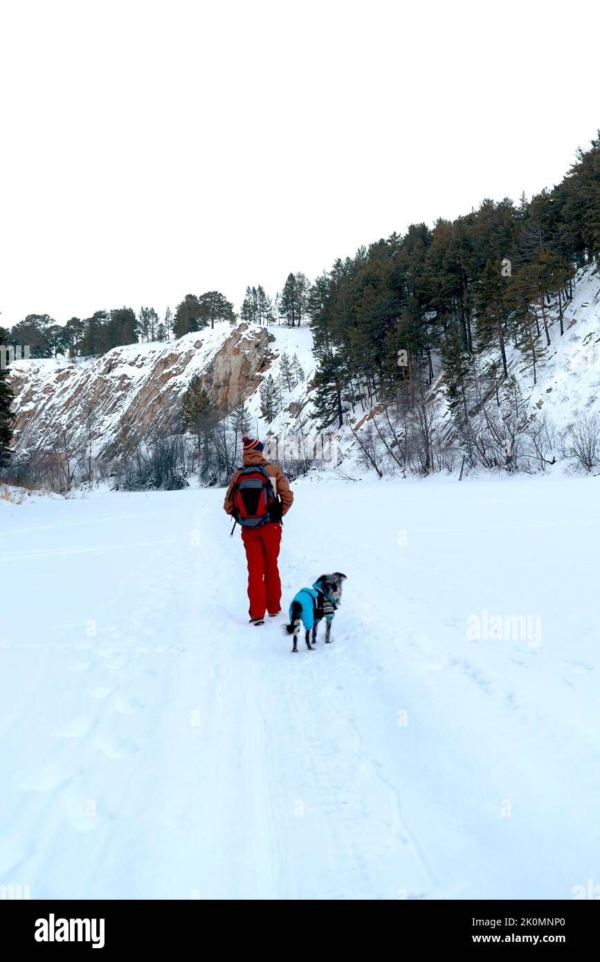 Vue arrière jeune homme en rouge brun chaud avec sac à dos avec chien mixte en costume bleu chaud marchant dans la neige parmi les rochers et les falaises en hiver AC Banque D'Images