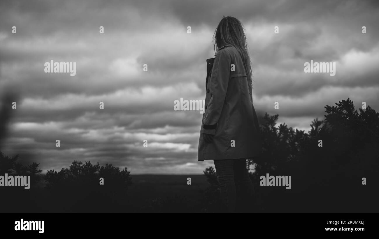 Un noir et blanc d'une femme regardant dans la distance capturée de derrière sous un ciel nuageux Banque D'Images