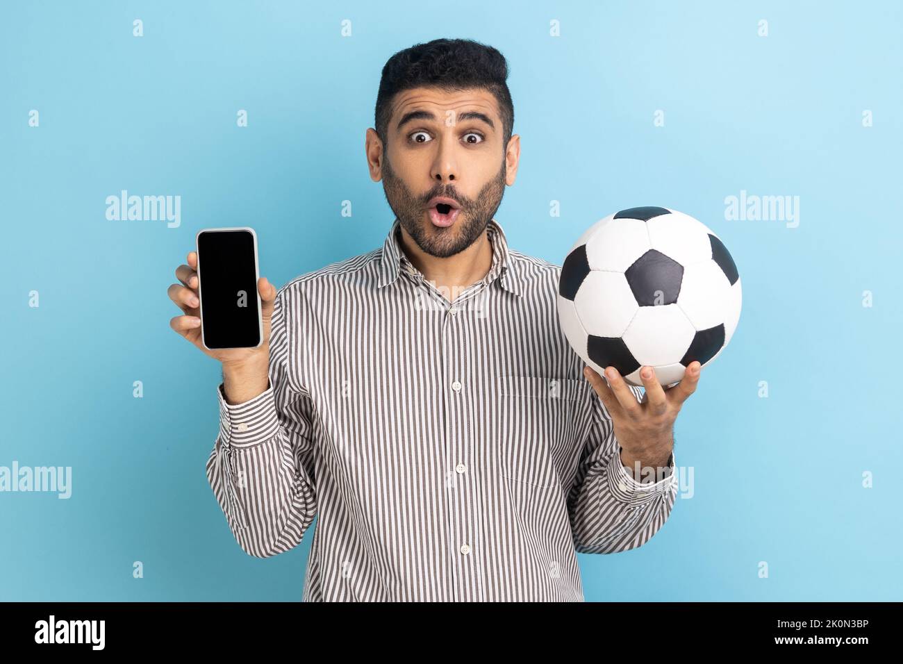 Surpris barbu homme tenant le ballon de football et smartphone vide écran noir, choqué par la facilité de réservation de billet de championnat, portant une chemise rayée. Studio d'intérieur isolé sur fond bleu. Banque D'Images
