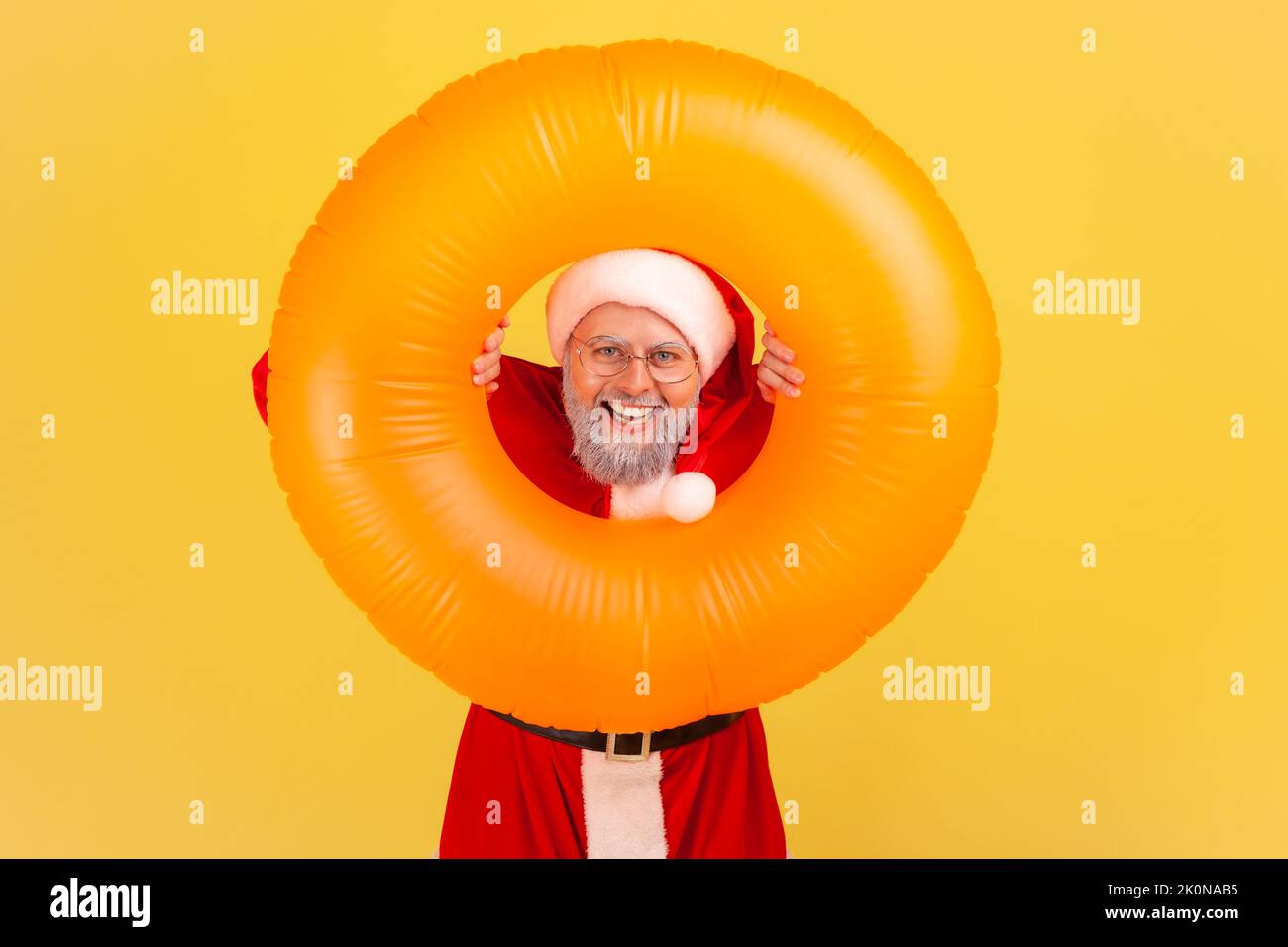 Homme âgé souriant avec barbe grise en costume du père noël tenant l'anneau en caoutchouc orange entre les mains, regardant la caméra avec une expression heureuse, visite d'hiver. Studio d'intérieur isolé sur fond jaune. Banque D'Images