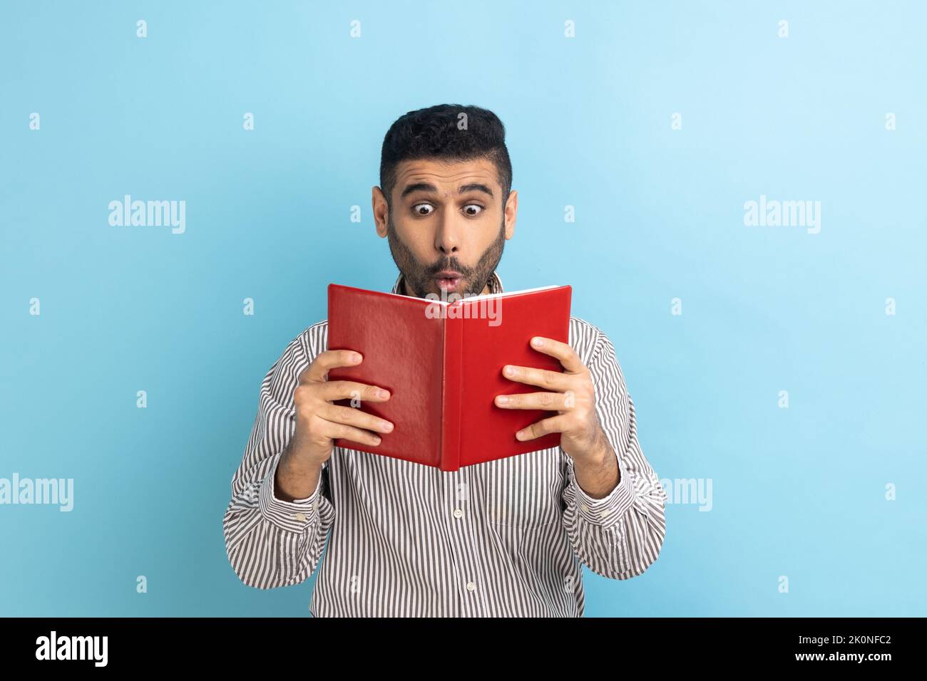 Jeune adulte choqué étonnant homme d'affaires lisant le livre, lisant avec de grands yeux, se tenant absorbé par l'intrigue stupéfaite, portant une chemise rayée. Studio d'intérieur isolé sur fond bleu. Banque D'Images Jeune adulte choqué étonnant homme d'affaires lisant le livre, lisant avec de grands yeux, se tenant absorbé par l'intrigue stupéfaite, portant une chemise rayée. Studio d'intérieur isolé sur fond bleu. Banque D'Images