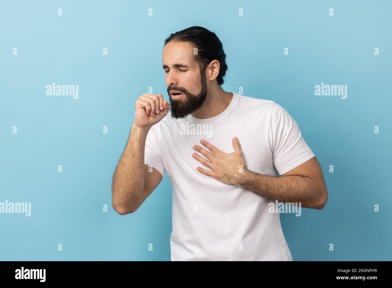 Portrait d'un malade malsain avec une barbe et des cheveux recueillis portant un T-shirt blanc ayant des symptômes de grippe, toux, gardant la main près de la bouche. Studio d'intérieur isolé sur fond bleu. Banque D'Images