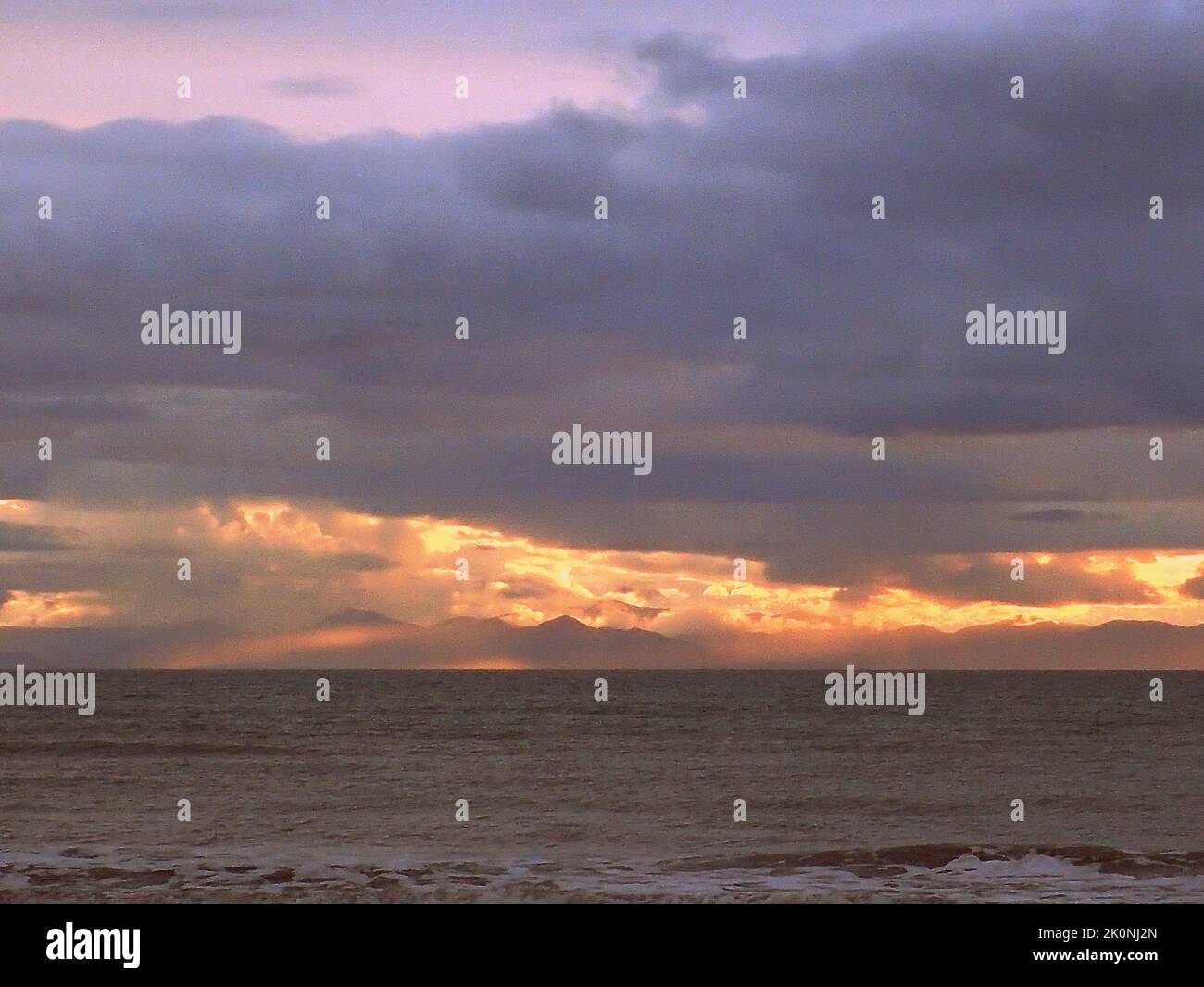 Les rayons du coucher du soleil traversent des nuages bas au-dessus de l'île du Sud, en Nouvelle-Zélande, vu de la plage de Raumati sur la partie inférieure de l'île du Nord Banque D'Images