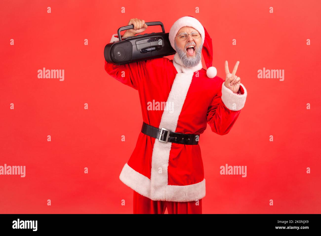 Positif drôle homme âgé avec barbe grise portant le costume du père noël debout et tenant le magnétophone montrant le signe v, célébrant la fête de Noël. Studio d'intérieur isolé sur fond rouge. Banque D'Images