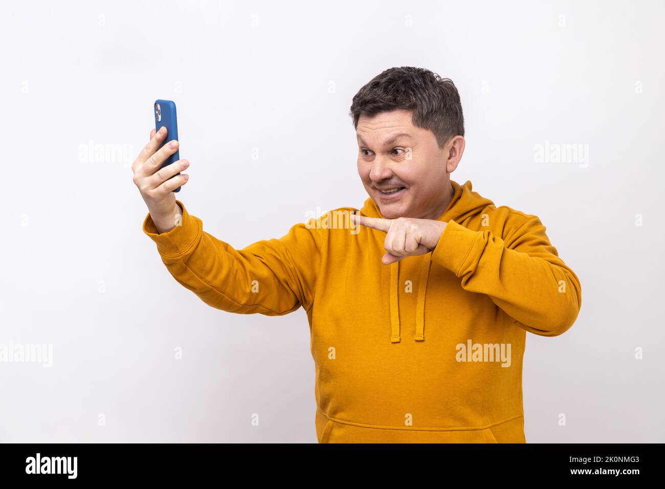 Portrait d'un homme d'âge moyen souriant pointant vers l'appareil photo du téléphone portable lors d'un appel vidéo ou d'une diffusion en direct, portant un sweat à capuche de style urbain. Studio d'intérieur isolé sur fond blanc. Banque D'Images