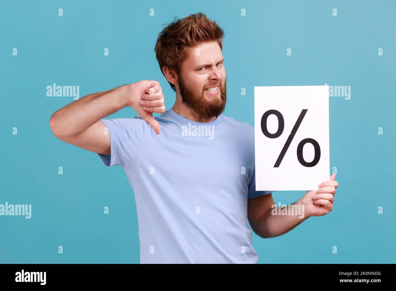 Portrait d'un jeune homme barbu adulte de belle taille tenant le papier avec le signe de pourcentage, regardant l'appareil photo avec le visage frognant et montrant le pouce vers le bas. Studio d'intérieur isolé sur fond bleu. Banque D'Images