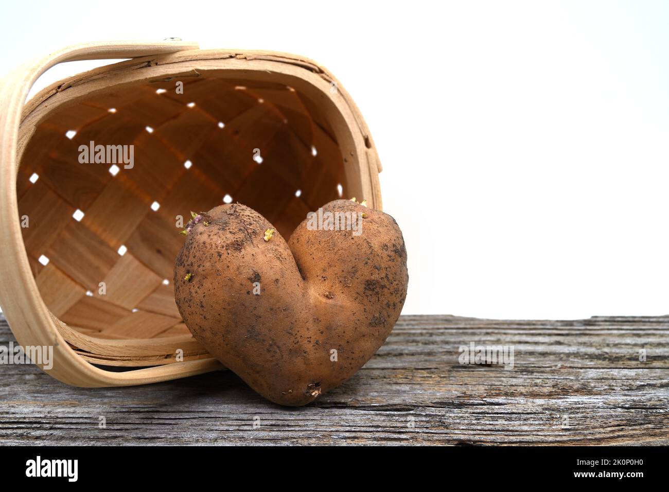 Pomme de terre en forme de coeur contre un panier tissé sur une table en bois abîmé sur un blanc Banque D'Images