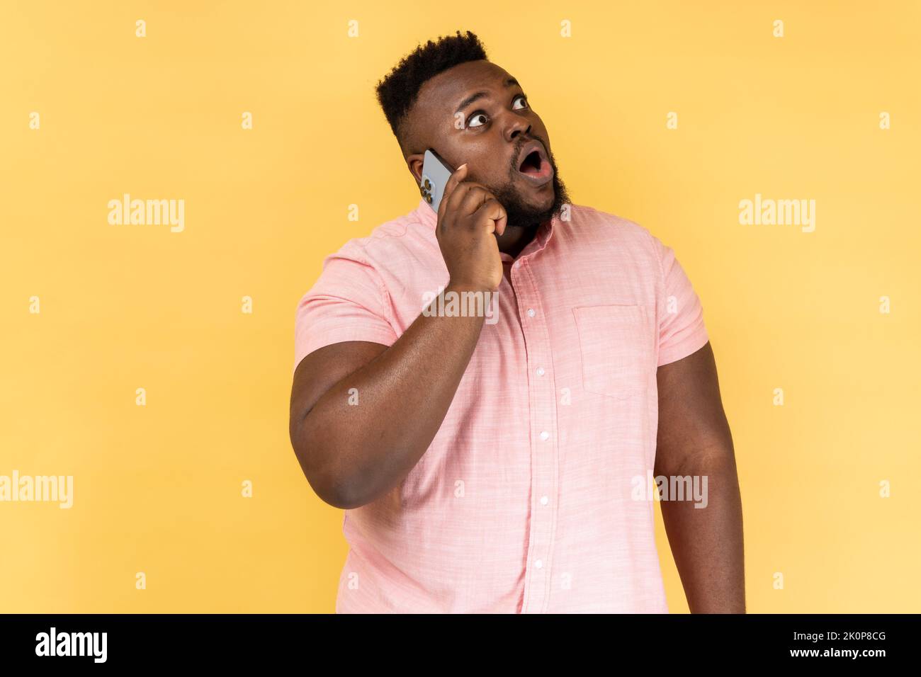 Portrait d'un homme choqué portant une chemise rose discutant avec un ami sur un téléphone portable, regardant loin avec surpris étonné, entendre des nouvelles de rupture. Studio d'intérieur isolé sur fond jaune. Banque D'Images