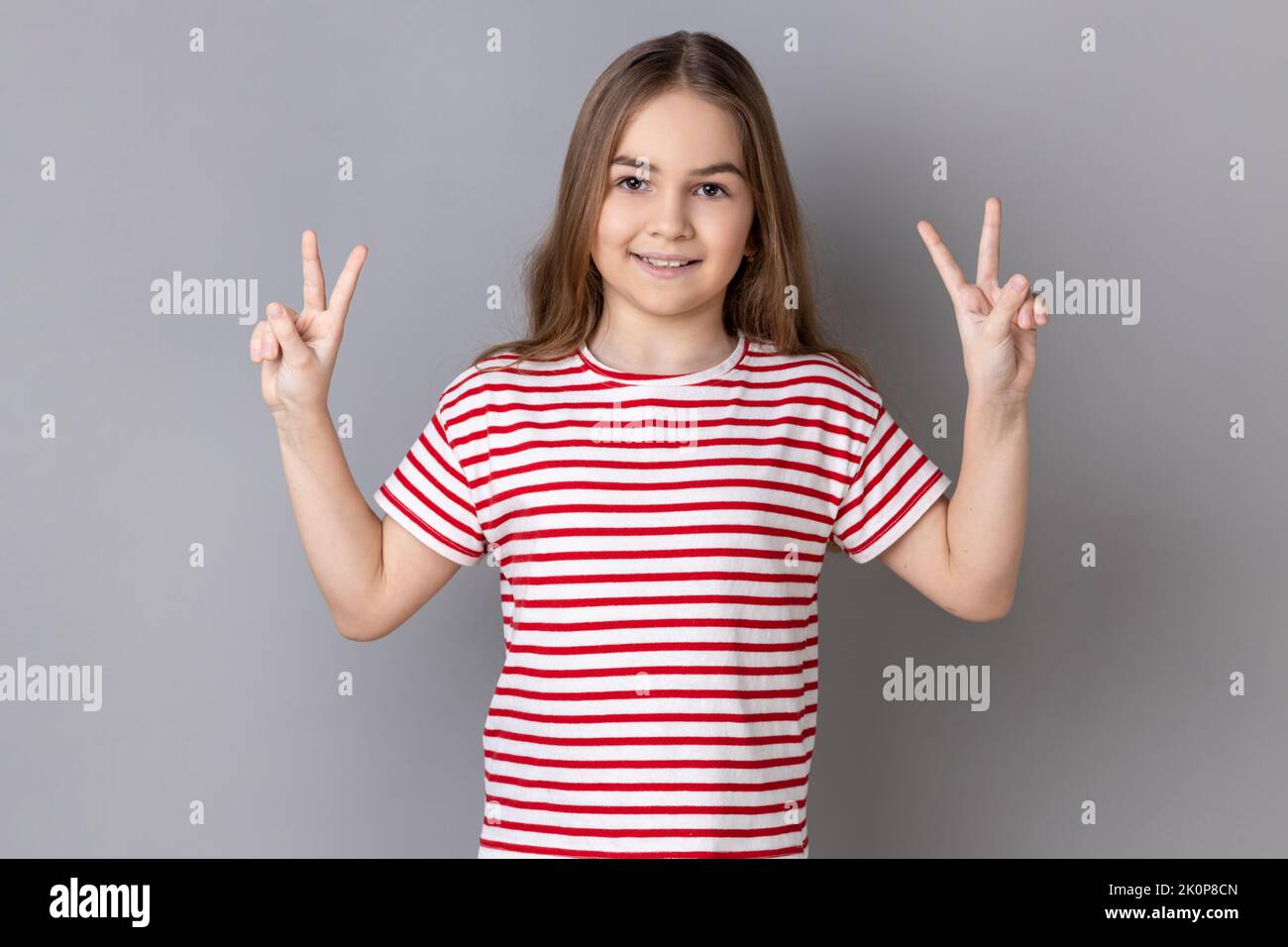 Portrait de positive optimiste adorable petite fille portant un T-shirt rayé gestante signe de victoire à la caméra, le succès ou la réalisation. Prise de vue en studio isolée sur fond gris. Banque D'Images