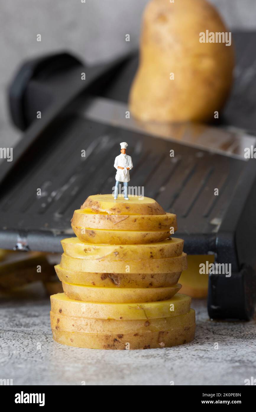 Petit chef modèle échelle, debout sur des pommes de terre empilées, avec coupe-cuisine mandoline en arrière-plan Banque D'Images
