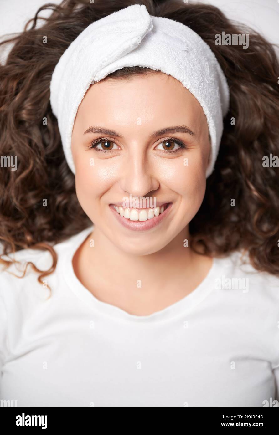 Belle jeune femme aux cheveux bouclés portant un bandeau blanc tout en regardant l'appareil photo et en souriant. Une femme gaie avec une peau parfaite exprimant des émotions positives. Banque D'Images