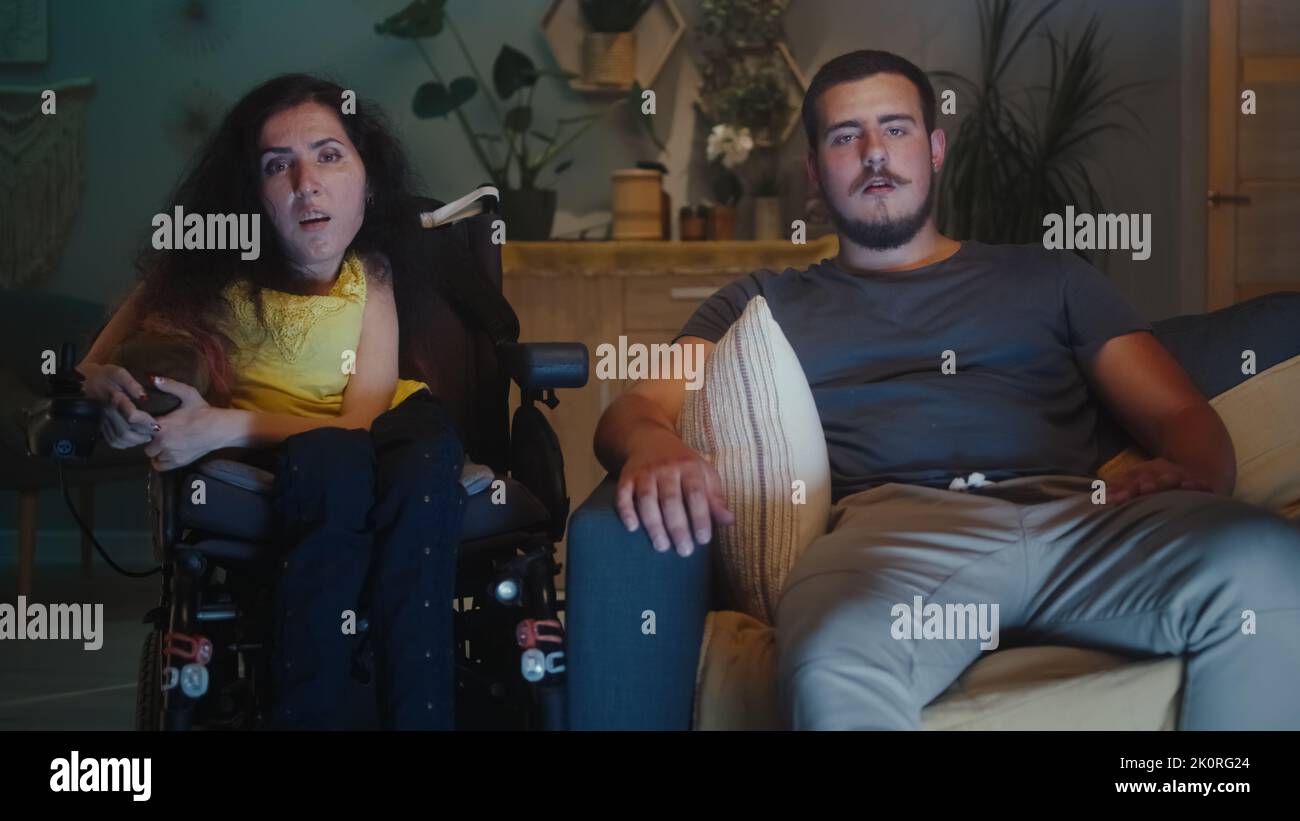 Femme atteinte d'atrophie spinale assise dans un fauteuil roulant à côté de son mari et ils regardent les nouvelles à la télévision le soir ensemble Banque D'Images