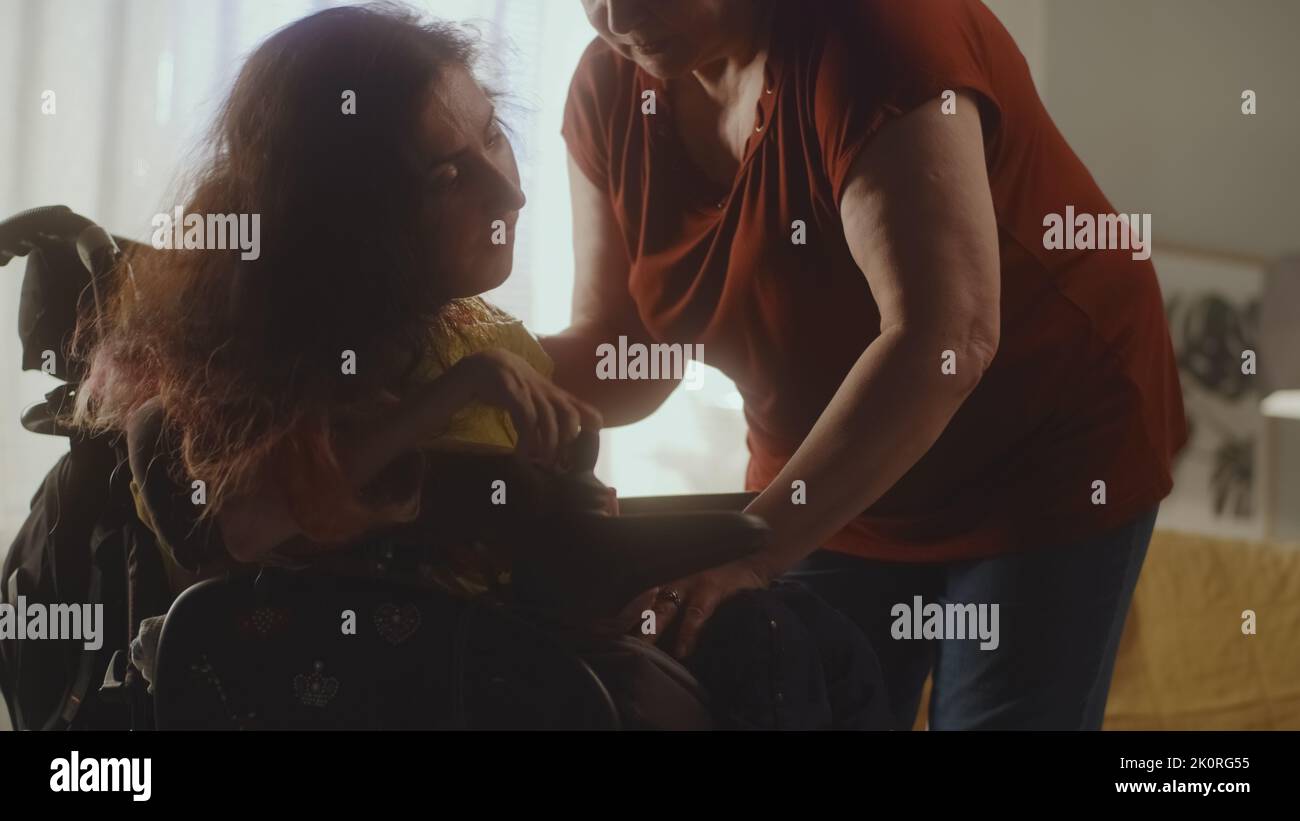 Femme âgée aidant la femme avec une atrophie musculaire spinale en mettant en place un fauteuil roulant motorisé moderne pour s'asseoir confortablement Banque D'Images