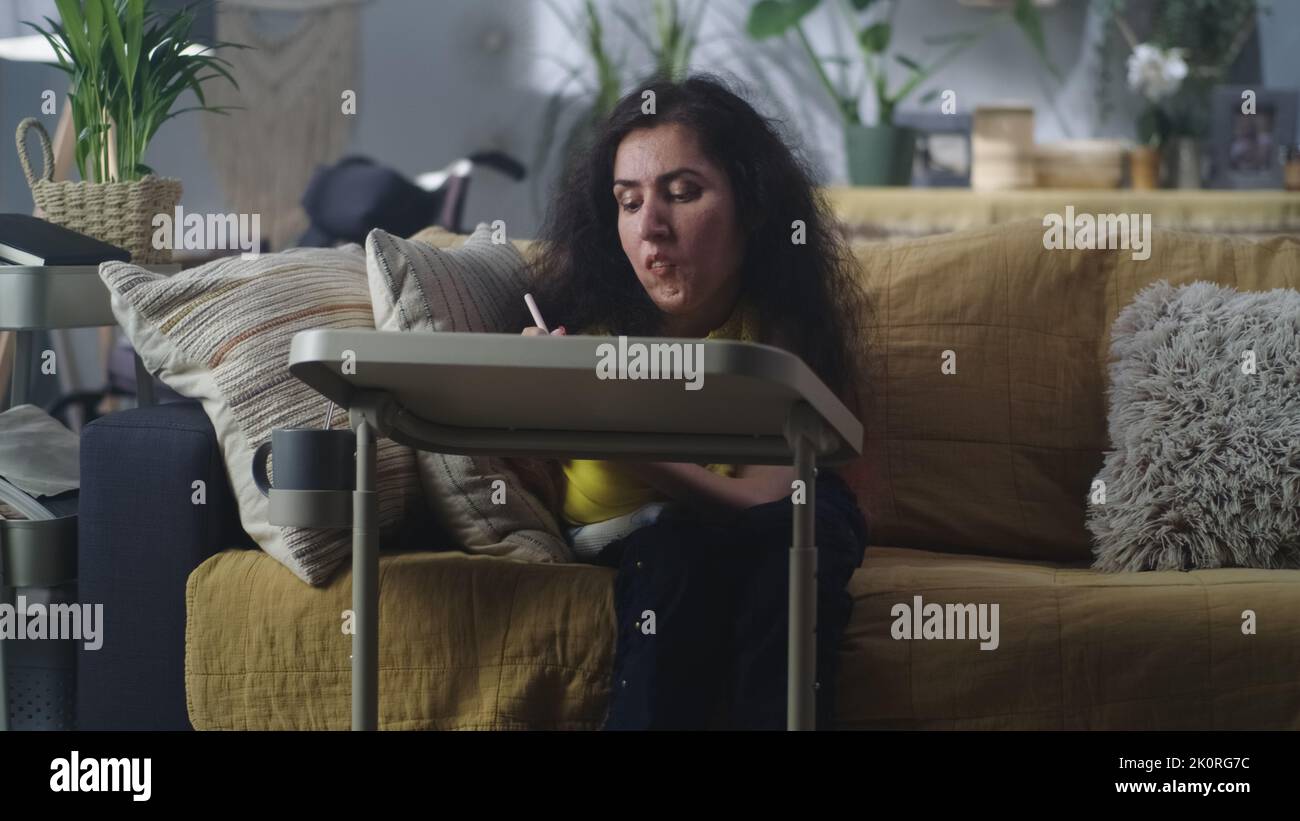Femme atteinte d'atrophie musculaire de la colonne vertébrale assise sur un canapé à la maison à la table et dessin avec un crayon numérique dans son temps libre Banque D'Images