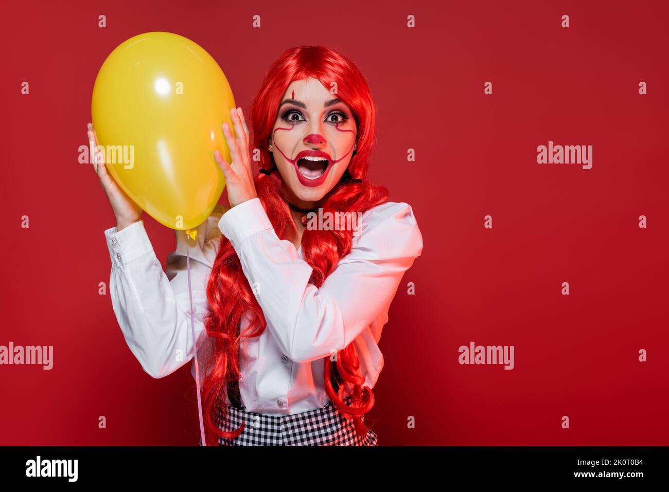 femme choquée avec le maquillage clown tenant le ballon jaune et criant isolé sur le rouge Banque D'Images