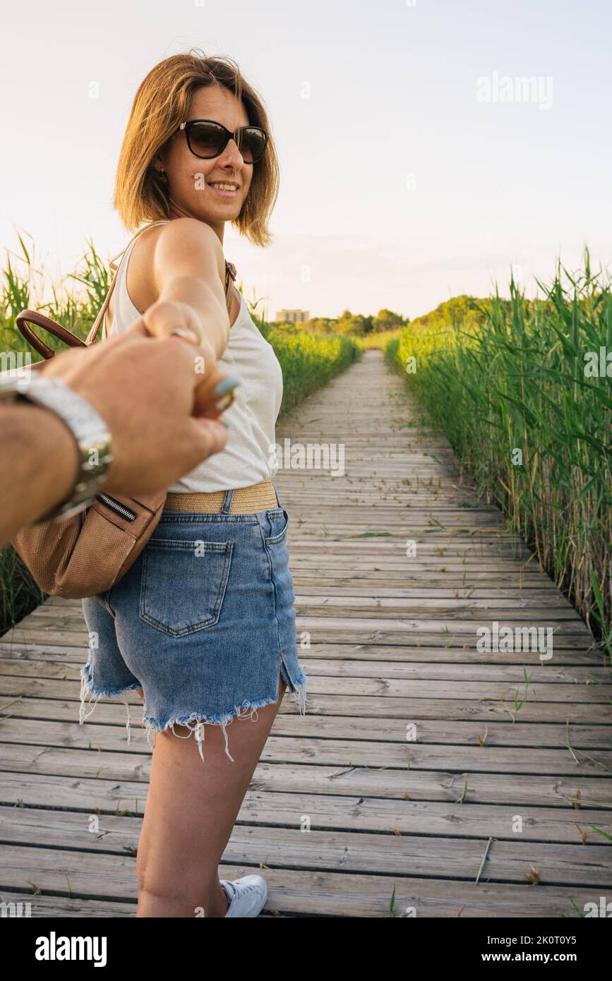 Femme habillée décontractée marchant dans la nature au coucher du soleil, tenant ma main dans la première personne et souriant. Vue verticale Banque D'Images