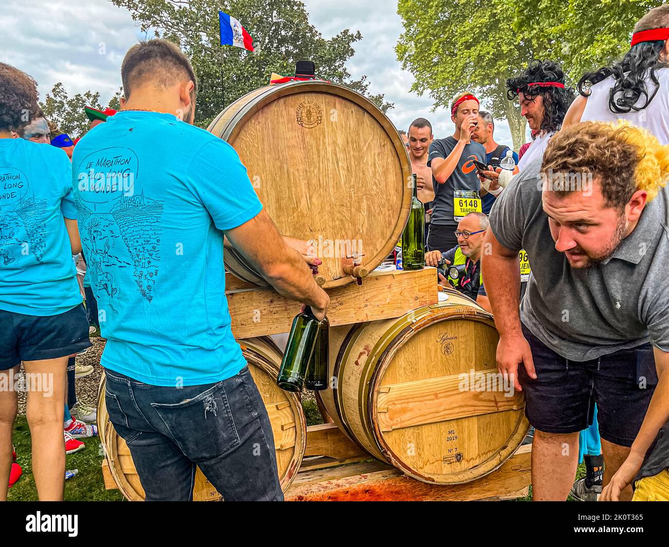 Au Château Larose-Trintaudon à Saint-Laurent-Médoc, en France, un assistant verse du vin rouge directement d'un tonneau barrique dans des verres pendant le 36e Marathon des Châteaux du Médoc – le millésime 2015 coule directement du robinet. Un coureur porte un T-shirt arborant l'inscription "Le Marathon le plus long du monde", un drapeau français flottant au-dessus de la scène. Banque D'Images
