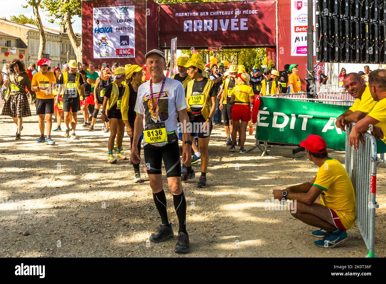 Le journaliste Georg Berg atteint l'arche d'arrivée "Arrivée" du 36e Marathon des Châteaux du Médoc à Pauillac, en France. Autour de lui, de nombreux coureurs portent des gilets de sécurité jaunes ou des pièces de costume jaunes. Son T-shirt représente le visage du personnage de télévision fictif Max Headroom. Banque D'Images