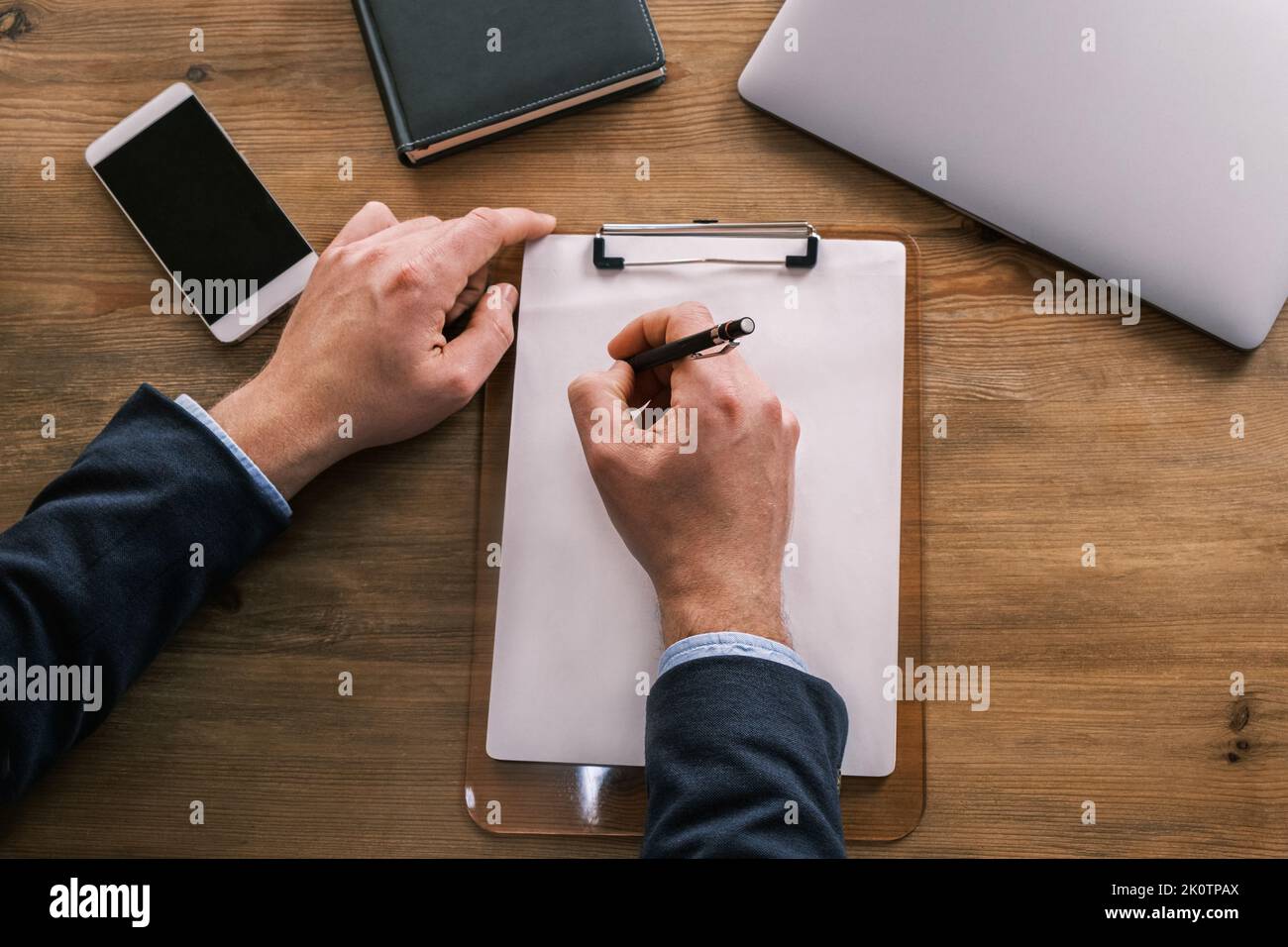 Vue de dessus des mains de sexe masculin écrivant quelque chose dans un papier à en-tête vierge attaché à un presse-papiers. Homme d'affaires travaillant à la table à côté de l'ordinateur portable, bloc-notes et Banque D'Images