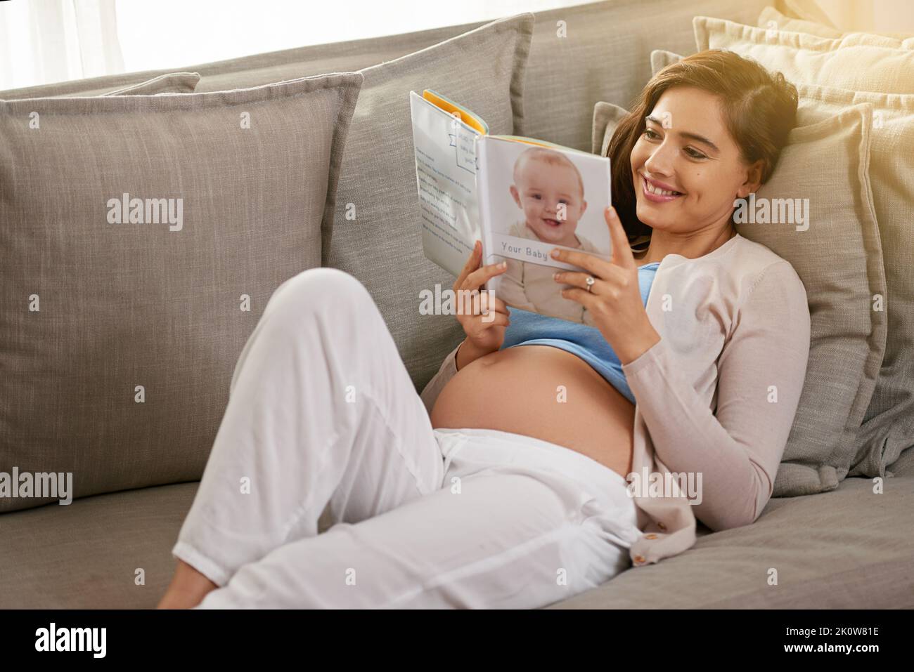 Ce livre répond à toutes mes questions de grossesse et de bébé. Une jeune femme enceinte lisant un livre de bébé à la maison. Banque D'Images