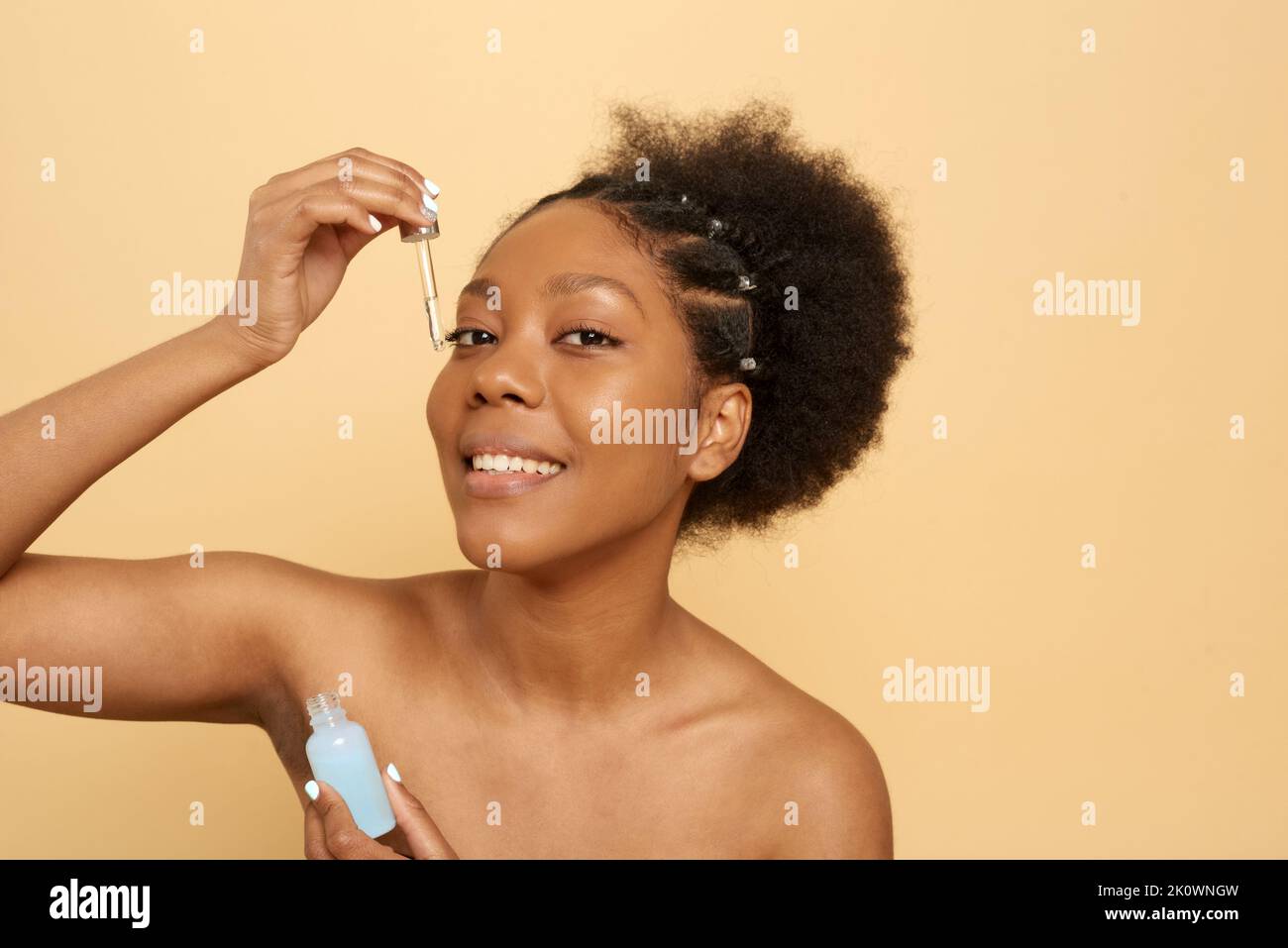 Le concept de la beauté, des cosmétiques et des soins de la peau. Portrait d'une jeune femme afro-américaine souriante aux épaules nues appliquant du sérum facial ou de l'oi Banque D'Images