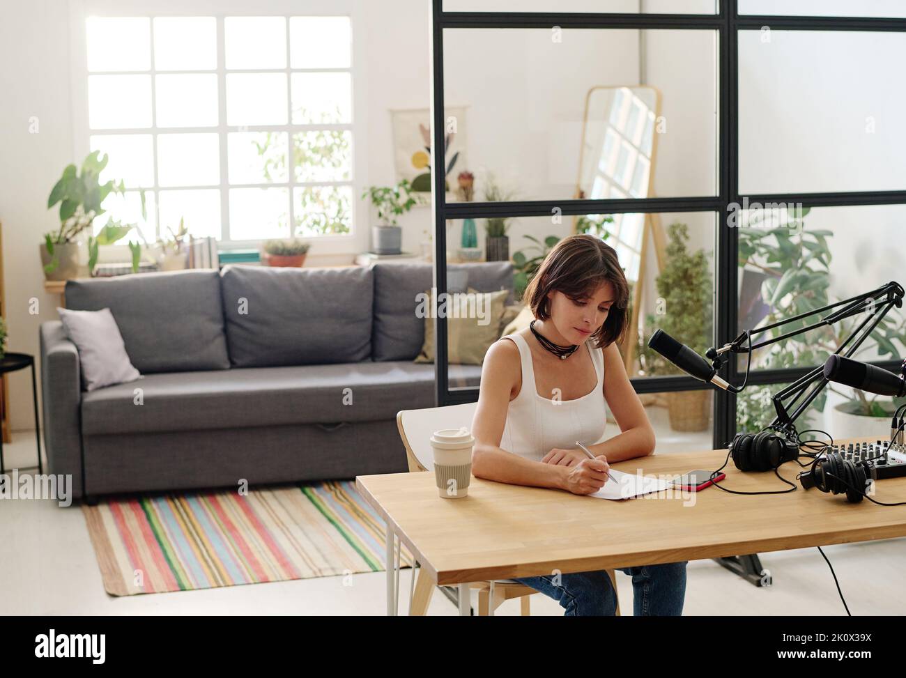 Jeune femme hôte de radio en tenue décontractée prenant des notes sur papier tout en étant assise sur le lieu de travail devant le microphone et la carte son Banque D'Images