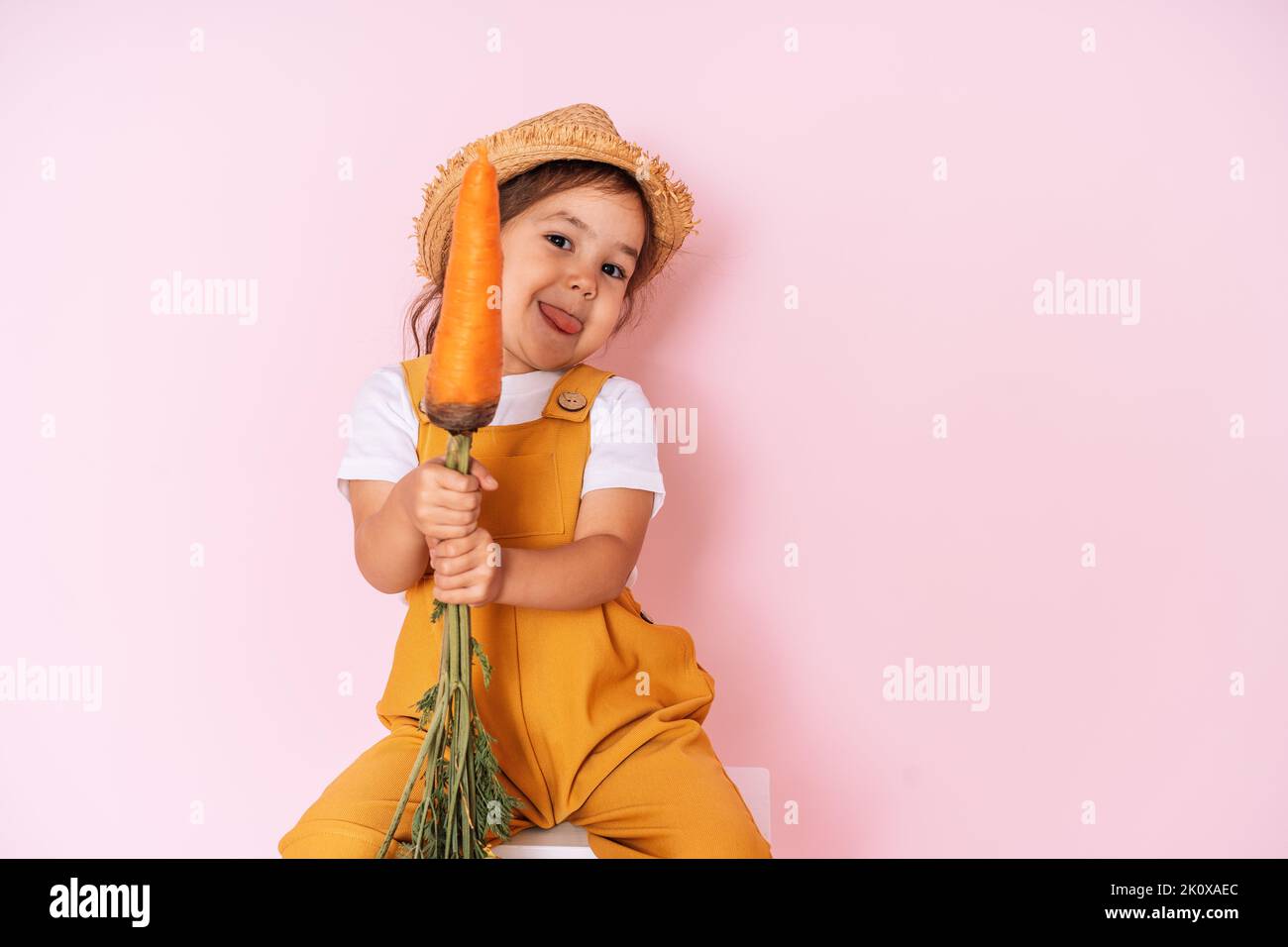 Petite fille dans une combinaison orange est assise sur un escabeau. Enfant tenant la carotte devant un fond rose Banque D'Images