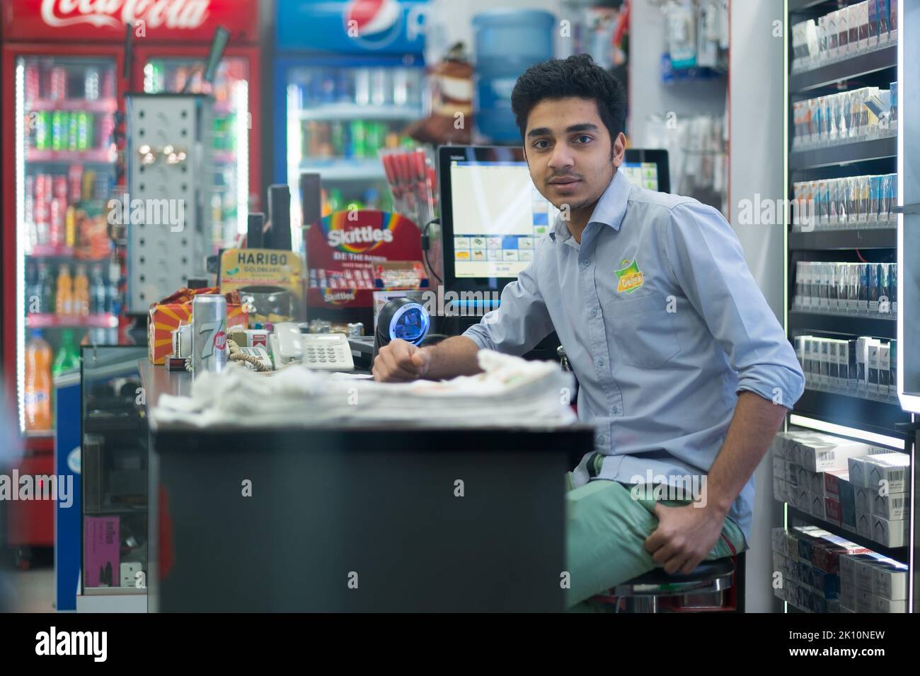 Jeune caissière indienne dans un magasin de proximité à Dubaï, Émirats arabes Unis. L'homme est assis avec un bras sur le comptoir et regarde la caméra. Banque D'Images