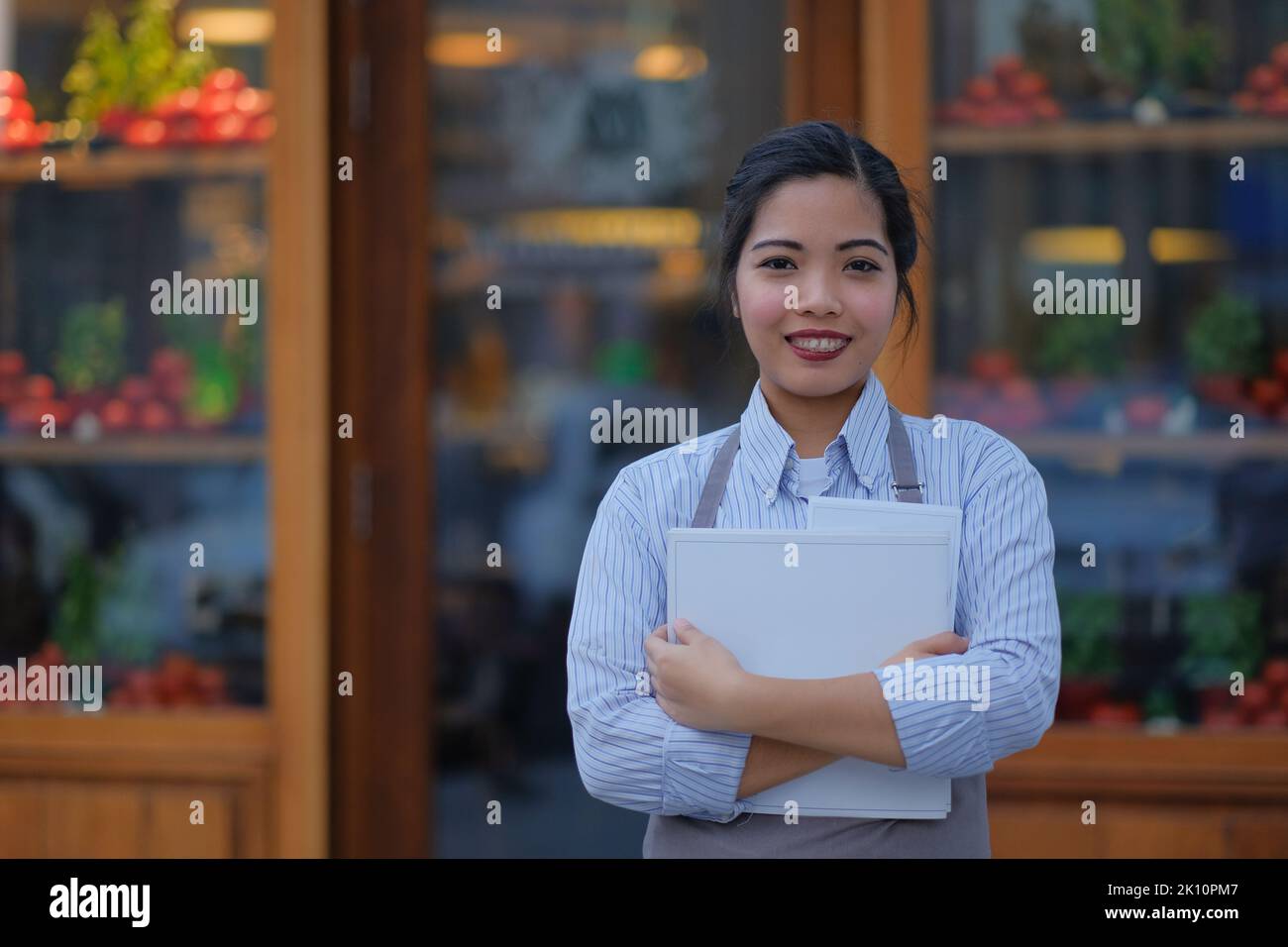 Une jeune serveuse philippine attend devant la fenêtre d'un restaurant italien avec des menus et un sourire. Portrait extérieur d'une femme asiatique dans l'industrie des services. Banque D'Images