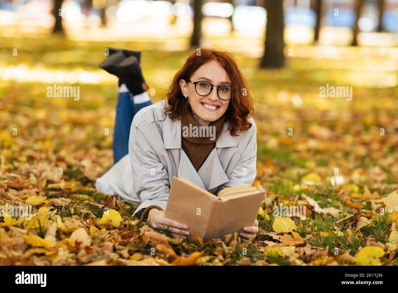 Bonne jolie femme caucasienne à cheveux rouges en imperméable et lunettes lit le livre se trouve sur terre dans le parc Banque D'Images