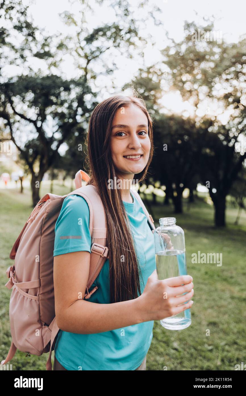 Adolescente portant une chemise turquoise et un sac à dos rose, tenant une bouteille de verre avec de l'eau dans la nature Banque D'Images