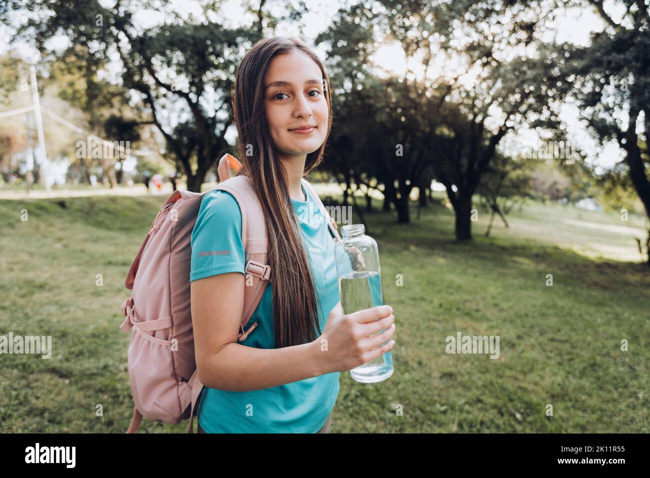 Adolescente portant une chemise turquoise et un sac à dos rose, tenant une bouteille de verre avec de l'eau dans la nature Banque D'Images