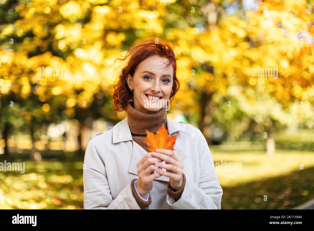 Bonne caucasienne femme à tête rouge millénaire en imperméable tenir la feuille jaune dans les mains, jouir de la liberté Banque D'Images