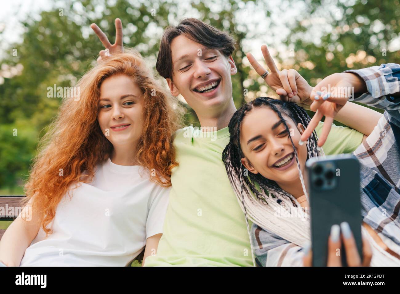 Vue de face de trois amis assis sur un banc en bois dans un parc prenant un selfie à l'aide d'un dispositif moderne. Selfie de jeunes souriants qui s'amusent Banque D'Images